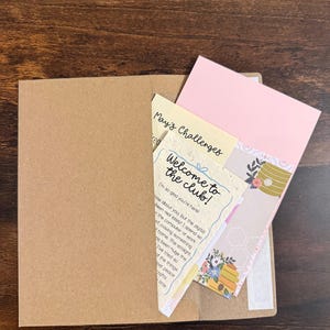 Pu&ograve; includere: Una busta in carta kraft marrone contenente diverse carte, tra cui una carta color crema con il testo "Welcome to the club!" e "May's Challenges", una carta rosa chiaro a tinta unita e una carta decorativa con un'illustrazione di un alveare e fiori.