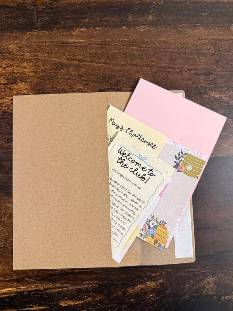 Pu&ograve; includere: Una busta in carta kraft marrone contenente diverse carte, tra cui una carta color crema con il testo "Welcome to the club!" e "May's Challenges", una carta rosa chiaro a tinta unita e una carta decorativa con un'illustrazione di un alveare e fiori.
