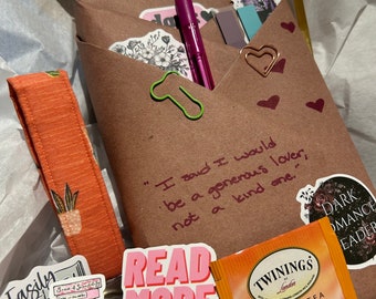May include: A brown paper package with a quote about being a generous lover, a pink pen, a green paper clip, and a sticker that says "Read More Books". There is also a box of Twinings Honeybush, Mandarin & Orange herbal tea.