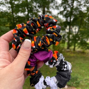 May include: A set of three Halloween-themed scrunchies. The scrunchies are made of black fabric with different patterns, including candy corn, polka dots, and a black and white pattern.