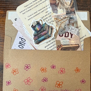 Pu&ograve; includere: Una busta di carta marrone decorata con fiori disegnati a mano in arancione e rosa e il testo "make today great!" con una faccina sorridente. All'interno ci sono vari articoli di carta, tra cui un adesivo di una tazza da t&egrave; su libri.