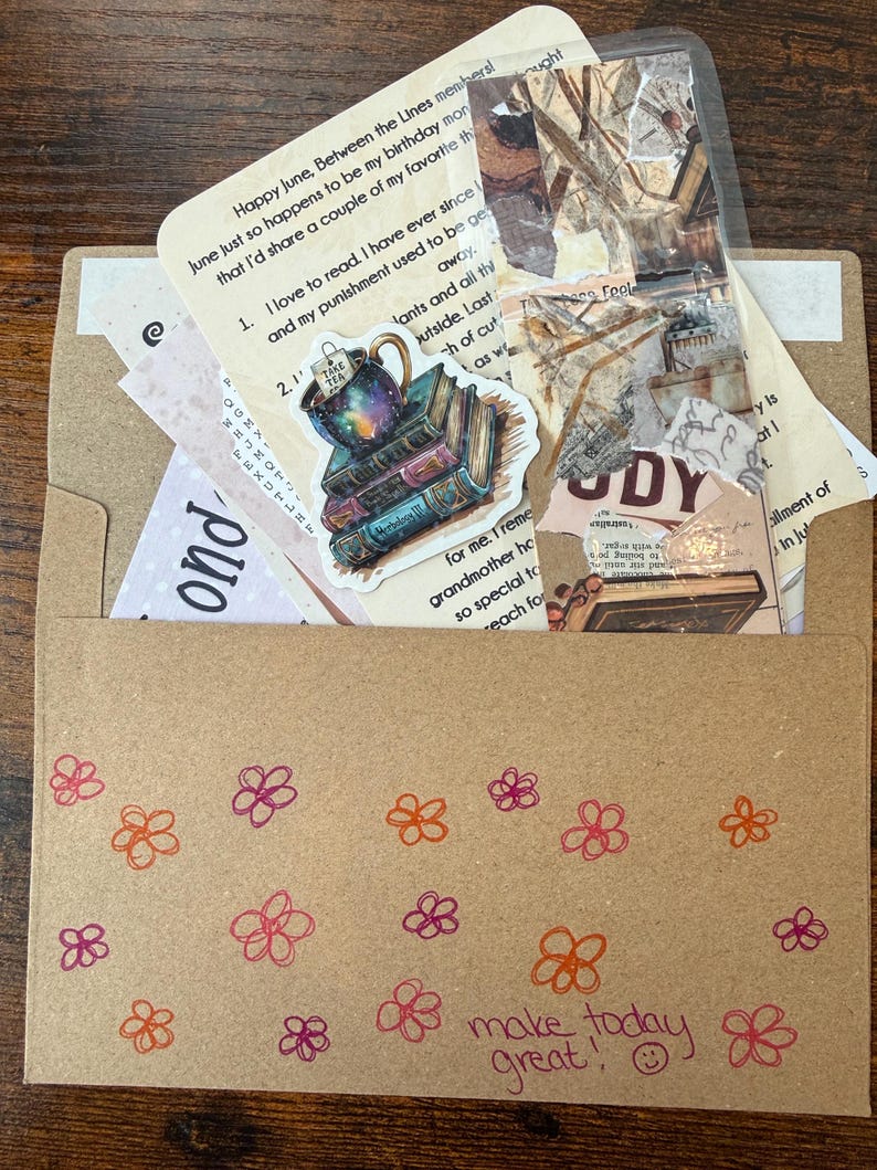 Pu&ograve; includere: Una busta di carta marrone decorata con fiori disegnati a mano in arancione e rosa e il testo "make today great!" con una faccina sorridente. All'interno ci sono vari articoli di carta, tra cui un adesivo di una tazza da t&egrave; su libri.