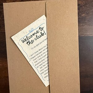 Pu&ograve; includere: Una carta color crema con una superficie ruvida e maculata inserita in una busta di carta kraft marrone. La carta presenta il testo blu scritto a mano Welcome to the club! e I'm so glad you're here, seguito da un paragrafo di testo nero.