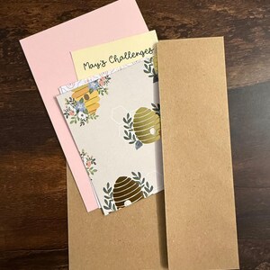 Pu&ograve; includere: Un set di cancelleria composto da una busta in carta kraft marrone, un cartoncino rosa chiaro, un cartoncino giallo pallido con la scritta "May's Challenges" in nero e un cartoncino bianco con un motivo ripetuto di alveari dorati e fogliame verde.
