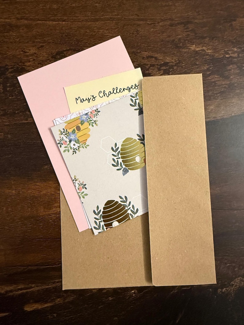 Pu&ograve; includere: Un set di cancelleria composto da una busta in carta kraft marrone, un cartoncino rosa chiaro, un cartoncino giallo pallido con la scritta "May's Challenges" in nero e un cartoncino bianco con un motivo ripetuto di alveari dorati e fogliame verde.