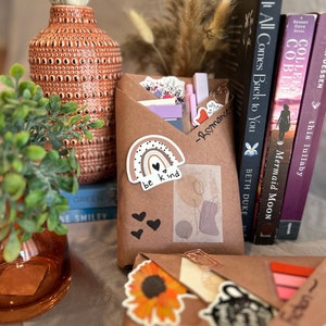 May include: A brown paper package with a rainbow sticker that says "be kind" and a few black heart stickers. The package is sitting on a table with a few books and a vase of dried flowers.