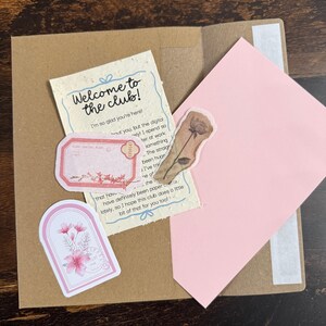 Pu&ograve; includere: Una busta di carta marrone contenente un biglietto color crema con il testo Welcome to the club! I'm so glad you're here!, un lembo di busta rosa, un'etichetta rettangolare rosa, un adesivo floreale ad arco e un adesivo con rosa marrone.