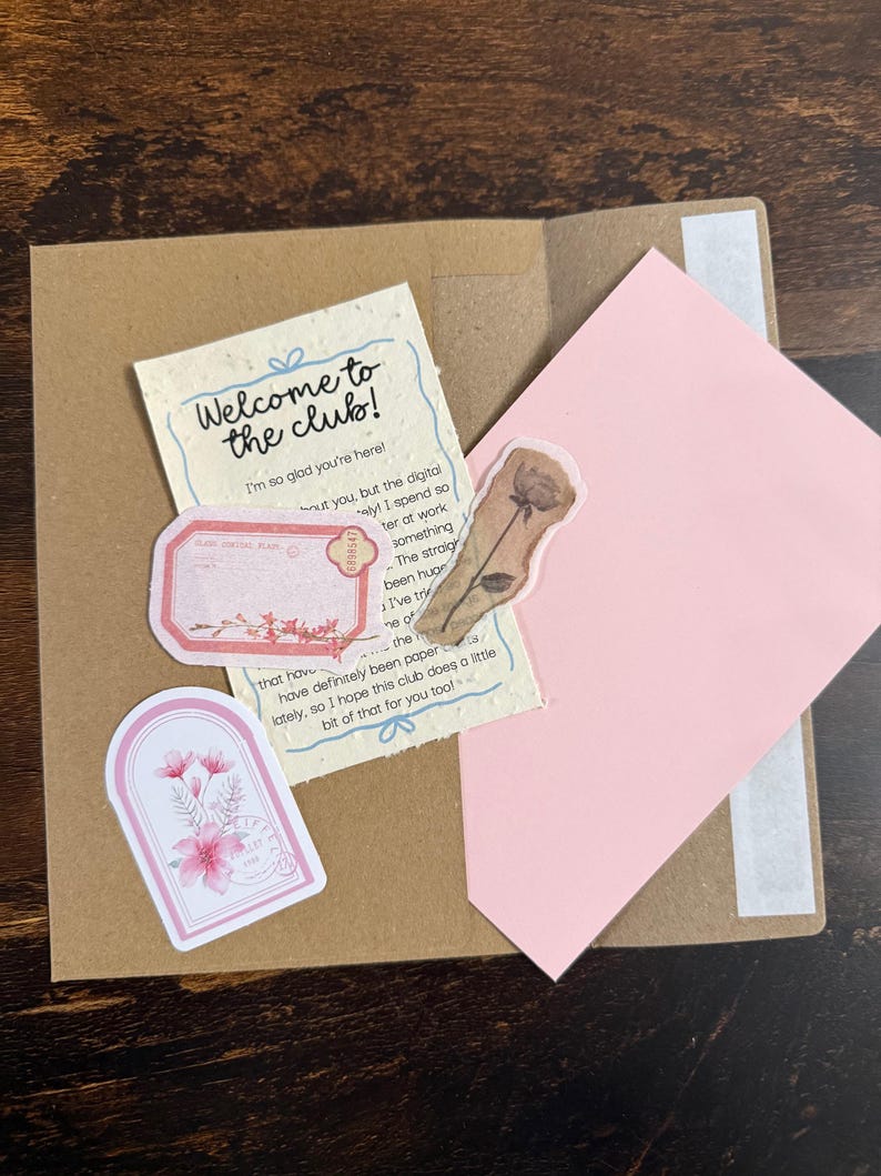 Pu&ograve; includere: Una busta di carta marrone contenente un biglietto color crema con il testo Welcome to the club! I'm so glad you're here!, un lembo di busta rosa, un'etichetta rettangolare rosa, un adesivo floreale ad arco e un adesivo con rosa marrone.