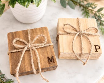 Puede incluir: Dos pilas de posavasos cuadrados de madera atados con lazos de cuerda de yute natural. Una pila es de madera marrón oscura con la letra M grabada, y la otra es de madera clara con la letra P grabada en la superficie del posavasos superior.