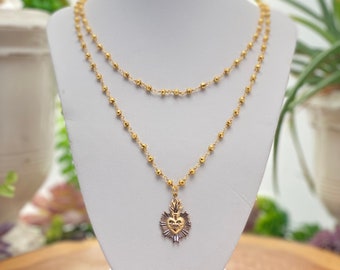 May include: A gold-toned necklace with a heart-shaped pendant. The pendant is surrounded by a decorative design and has a small flame on top. The necklace is displayed on a white mannequin.