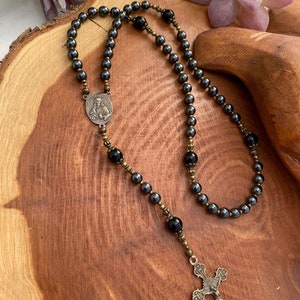 Peut inclure: Un chapelet en h&eacute;matite noire avec un pendentif argent&eacute; repr&eacute;sentant une figure religieuse et un charme en forme de croix. Le chapelet est pr&eacute;sent&eacute; sur une surface en bois.