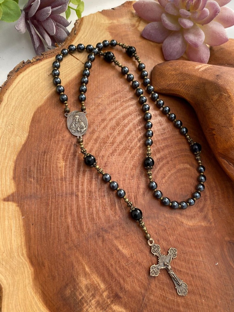 Peut inclure: Un chapelet en h&eacute;matite noire avec un pendentif argent&eacute; repr&eacute;sentant une figure religieuse et un charme en forme de croix. Le chapelet est pr&eacute;sent&eacute; sur une surface en bois.