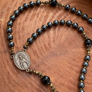 Peut inclure: Un chapelet en h&eacute;matite noire avec des accents dor&eacute;s. Le chapelet pr&eacute;sente un pendentif rond avec une repr&eacute;sentation de la Vierge Marie. Les perles sont entrecoup&eacute;es de perles d'onyx noir.