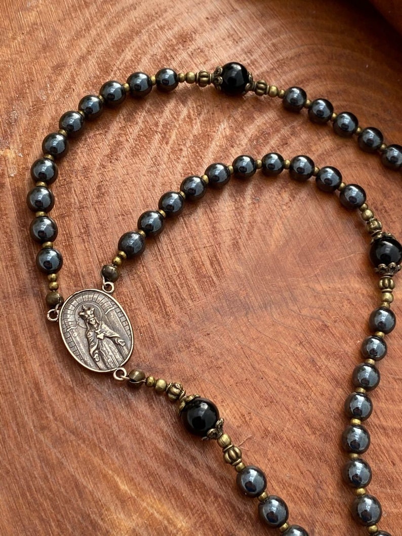 Peut inclure: Un chapelet en h&eacute;matite noire avec des accents dor&eacute;s. Le chapelet pr&eacute;sente un pendentif rond avec une repr&eacute;sentation de la Vierge Marie. Les perles sont entrecoup&eacute;es de perles d'onyx noir.