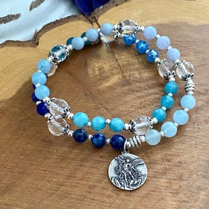 May include: A double-strand bracelet with blue and clear beads and a silver charm of an angel. The bracelet is on a wooden surface.