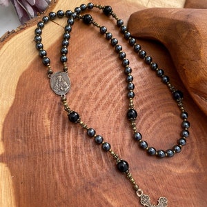 Peut inclure: Un chapelet en h&eacute;matite noire avec un centre en argent&eacute; repr&eacute;sentant une figure religieuse et un crucifix. Le chapelet est enfil&eacute;e avec des perles d'onyx noires et des accents en laiton.