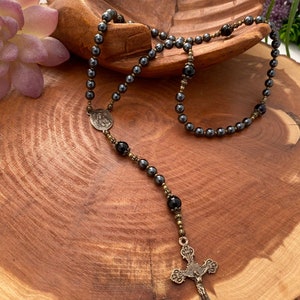Peut inclure: Un chapelet en h&eacute;matite noire avec un crucifix en bronze et une m&eacute;daille en bronze repr&eacute;sentant une figure religieuse. Le chapelet est pr&eacute;sent&eacute; sur une surface en bois.