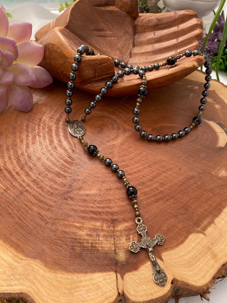 Peut inclure: Un chapelet en h&eacute;matite noire avec un crucifix en bronze et une m&eacute;daille en bronze repr&eacute;sentant une figure religieuse. Le chapelet est pr&eacute;sent&eacute; sur une surface en bois.