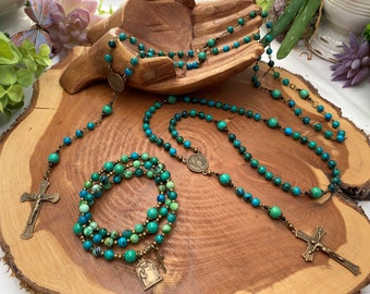 Peut inclure: Trois chapelets en turquoise et en pierre verte avec des accents bronze. Les chapelets sont disposés sur une surface en bois. Un chapelet est un bracelet et les deux autres sont des colliers. Chaque chapelet a une croix en bronze et une médaille en bronze.