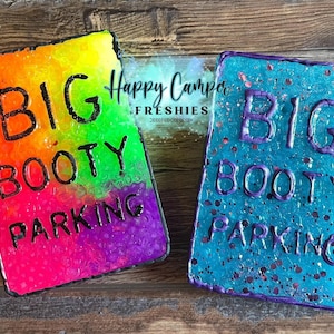 May include: Two rectangular air fresheners with the text "BIG BOOTY PARKING". One is a gradient of rainbow colors, the other is blue with glitter. The brand name "Happy Camper Freshies" is also visible.