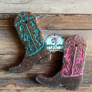May include: Two brown cowboy boot-shaped air fresheners. One boot has turquoise glitter accents and the other has pink glitter accents. The boots are on a wood background. The text "Happy Camper Freshies" is visible in the image.