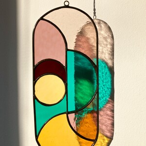 May include: A stained glass window hanging with a geometric design featuring pink, yellow, green, and brown glass pieces.
