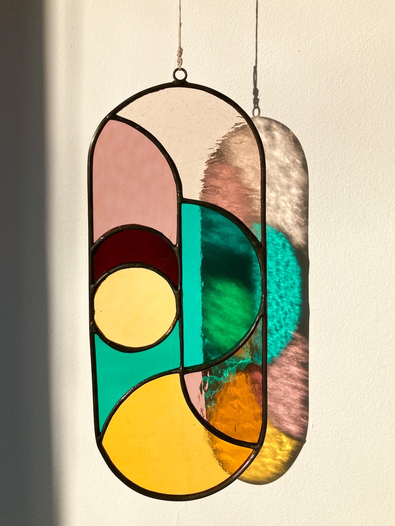 May include: A stained glass window hanging with a geometric design featuring pink, yellow, green, and brown glass pieces.
