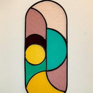 May include: A stained glass window hanging with a geometric design featuring pink, yellow, teal, and burgundy colors.