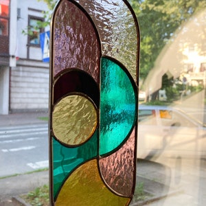May include: A stained glass window hanging features a geometric design with teal, gold, and purple glass pieces.