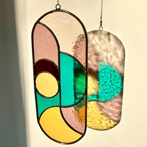 May include: A stained glass suncatcher with geometric shapes in shades of pink, yellow, green, and brown. The suncatcher is hanging from a white wall and casts a shadow on the wall.