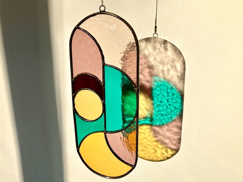 May include: A stained glass suncatcher with geometric shapes in shades of pink, yellow, green, and brown. The suncatcher is hanging from a white wall and casts a shadow on the wall.