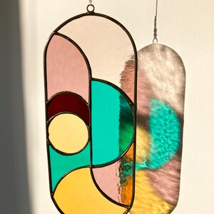 May include: A stained glass suncatcher with geometric shapes in shades of pink, yellow, red, and turquoise. The suncatcher is hanging from a string and casts a colorful shadow on the wall.