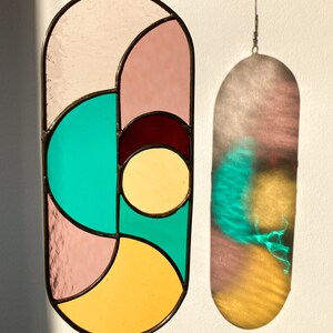 May include: A stained glass window hanging with a geometric design featuring pink, yellow, teal, and burgundy glass.