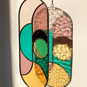 May include: A stained glass suncatcher with geometric shapes in shades of pink, green, and gold. The suncatcher is oval-shaped and hangs from a string.