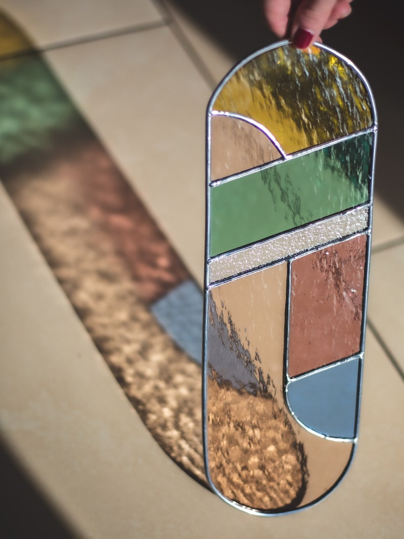 May include: A stained glass panel with geometric shapes in shades of brown, green, blue, and yellow. The panel is oval-shaped and has a silver frame.