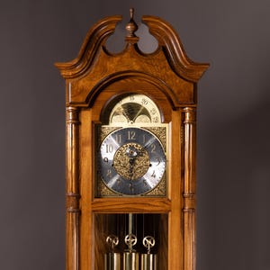 Puede incluir: Un reloj de pie alto de madera con acabado marrón claro. El reloj presenta una esfera cuadrada con números romanos, un péndulo y tres pesas. El reloj está encerrado en un mueble de madera con tallas decorativas, midiendo aproximadamente 180 cm de altura.