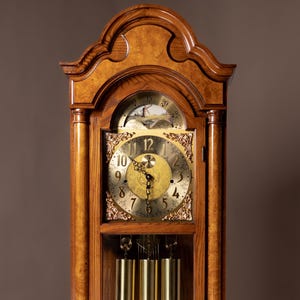 May include: A tall, wooden grandfather clock with a light brown finish. The clock features a gold-colored face with black Roman numerals, a pendulum, and weights. The clock is encased in a wooden cabinet with a glass door.