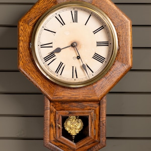 May include: Vintage wooden wall clock with a circular face, black Roman numerals on a cream backdrop. The clock has a brass bezel and a decorative pendulum visible through a small window.