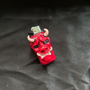 May include: A red lighter with a devil face design. The lighter features a sculpted face with horns, eyes, and a mouth. The lighter is a standard size and shape.