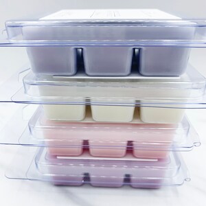 May include: A stack of four clear plastic containers with six individual wax melts in each. The wax melts are in different colors, including purple, white, and pink.