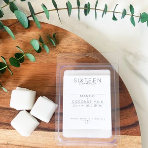 May include: A clear plastic container with a white wax melt inside. The wax melt is labeled "Mango + Coconut Milk" and is handcrafted in California. The container is sitting on a wooden surface with green eucalyptus leaves.