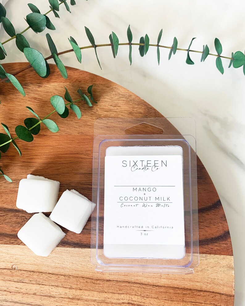 May include: A clear plastic container with a white wax melt inside. The wax melt is labeled "Mango + Coconut Milk" and is handcrafted in California. The container is sitting on a wooden surface with green eucalyptus leaves.