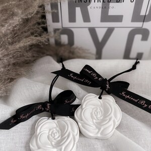 May include: Two white ceramic rose-shaped air fresheners with black ribbon tied around them. The ribbon has the text "Inspired By C" printed on it in white.