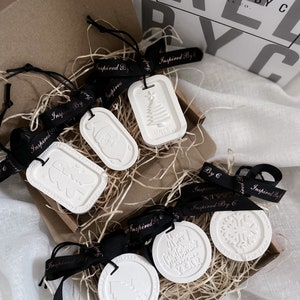 May include: A selection of white, embossed soy wax ornaments with black ribbon, presented in a brown box with straw. The ornaments display Christmas-themed designs and text, including a Christmas tree, Santa, and snowflake.
