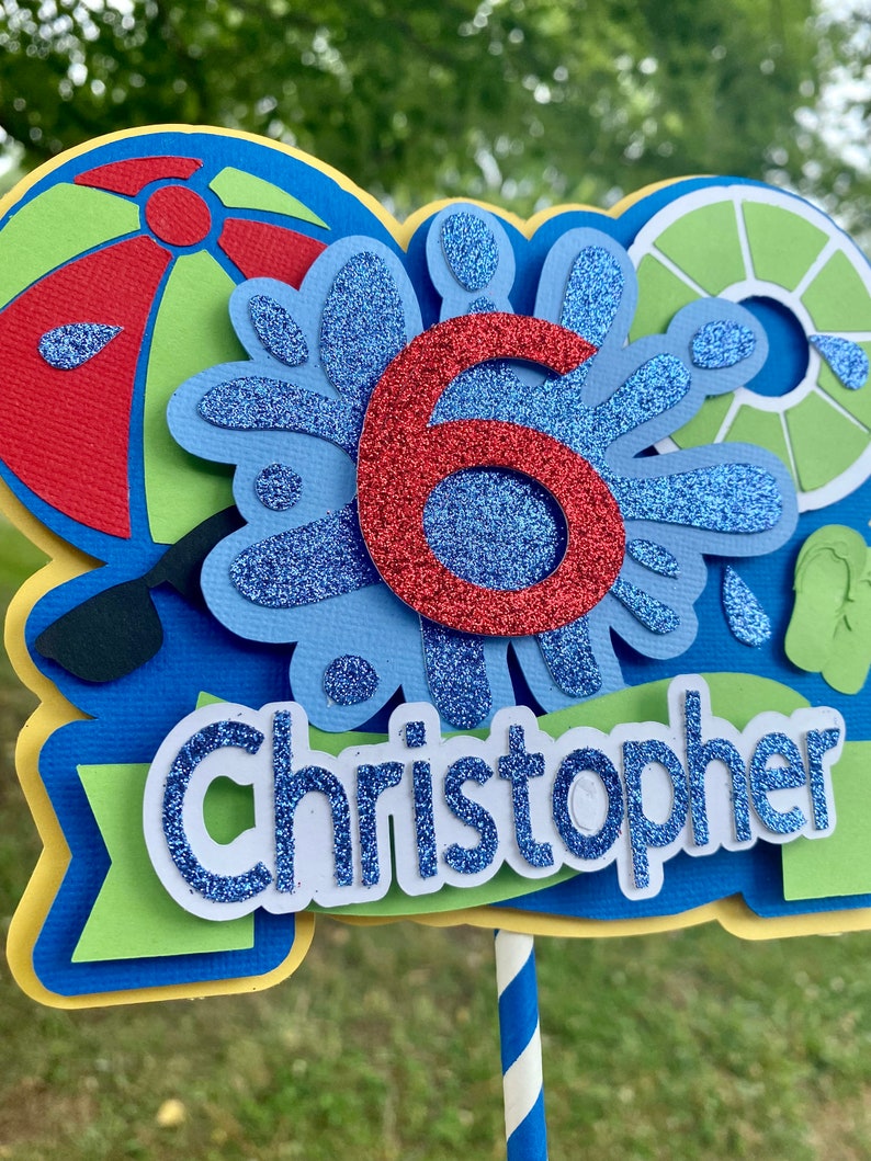 May include: A blue and green cake topper with a red number six, a blue and white splash design, a red and green beach ball, a green and white life preserver, and the name "Christopher" in blue glitter letters.