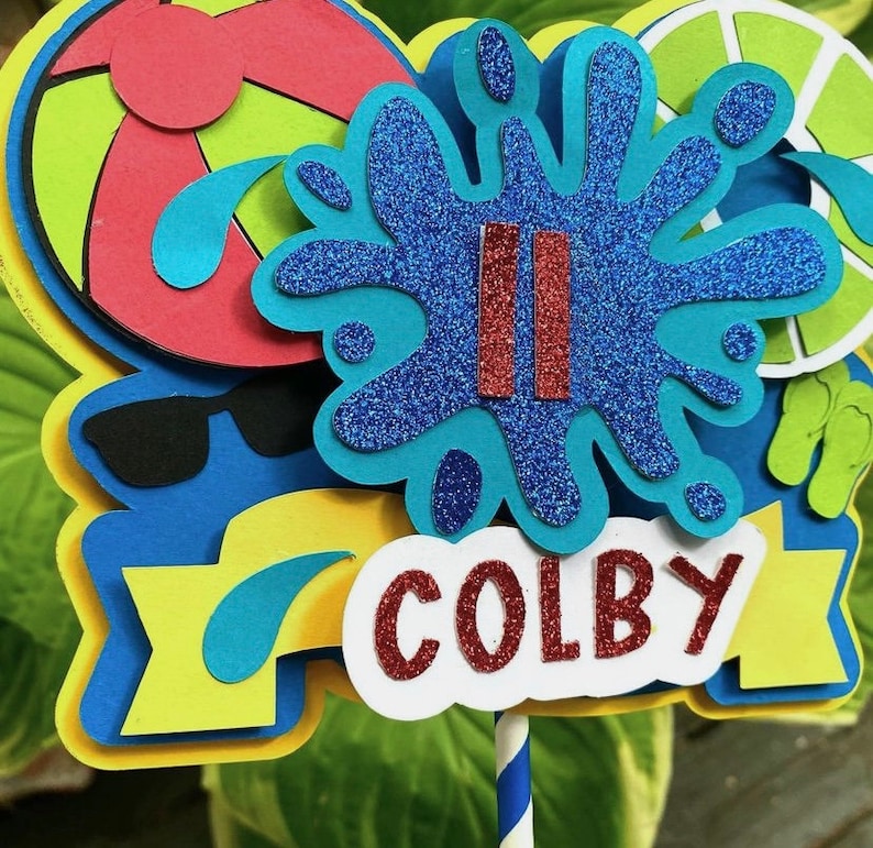 May include: A blue and white cake topper with a blue water splash design, red stripes, and the name "COLBY" in red letters. The topper is decorated with a yellow and blue banner, a red and white beach ball, and sunglasses.