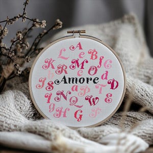 May include: A white embroidery hoop with pink and red letters spelling out the alphabet and the word "Amore".