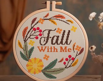 May include: A beige embroidery hoop with a cross-stitch design featuring a wreath of fall leaves, flowers, and berries. The text "Fall With Me" is stitched in brown thread.