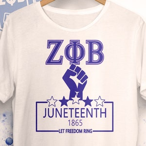 Könnte beinhalten: Weißes T-Shirt mit einem violetten griechischen Buchstaben-Design, das "ZΦΒ" und eine geballte Faust zeigt. Unter der Faust befindet sich ein Rechteck mit dem Text "JUNETEENTH 1865" und "LET FREEDOM RING". Der untere Teil des Shirts trägt den Text "RUN WITH THE YARD" in Blau.