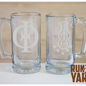 May include: Two clear glass beer mugs with handles. One mug features a large, etched Greek letter design, while the other has a hand gesture and Greek letters. The phrase "Run With The Yard" is printed in brown.